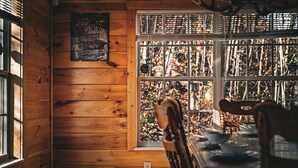 Interior de una cabaña de madera con una mesa de comedor, sillas y una ventana con vistas a árboles.
