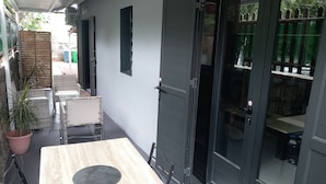 Un estrecho pasillo al aire libre con una mesa y sillas de madera, una planta en maceta y una puerta que da a una habitación con estanterías.