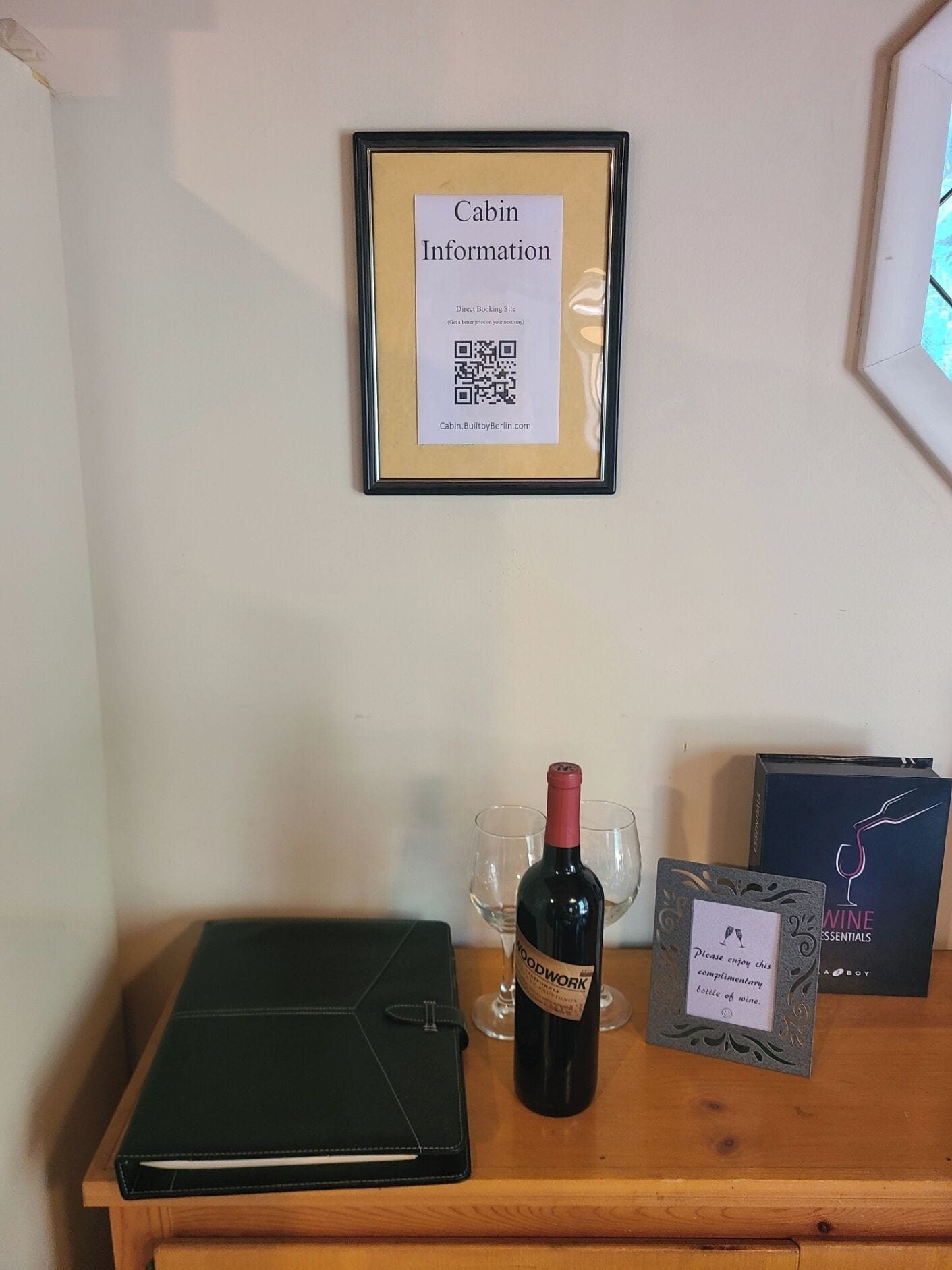 A framed sign with cabin information, a bottle of wine, glasses, a notebook, and a wine box on a wooden table.