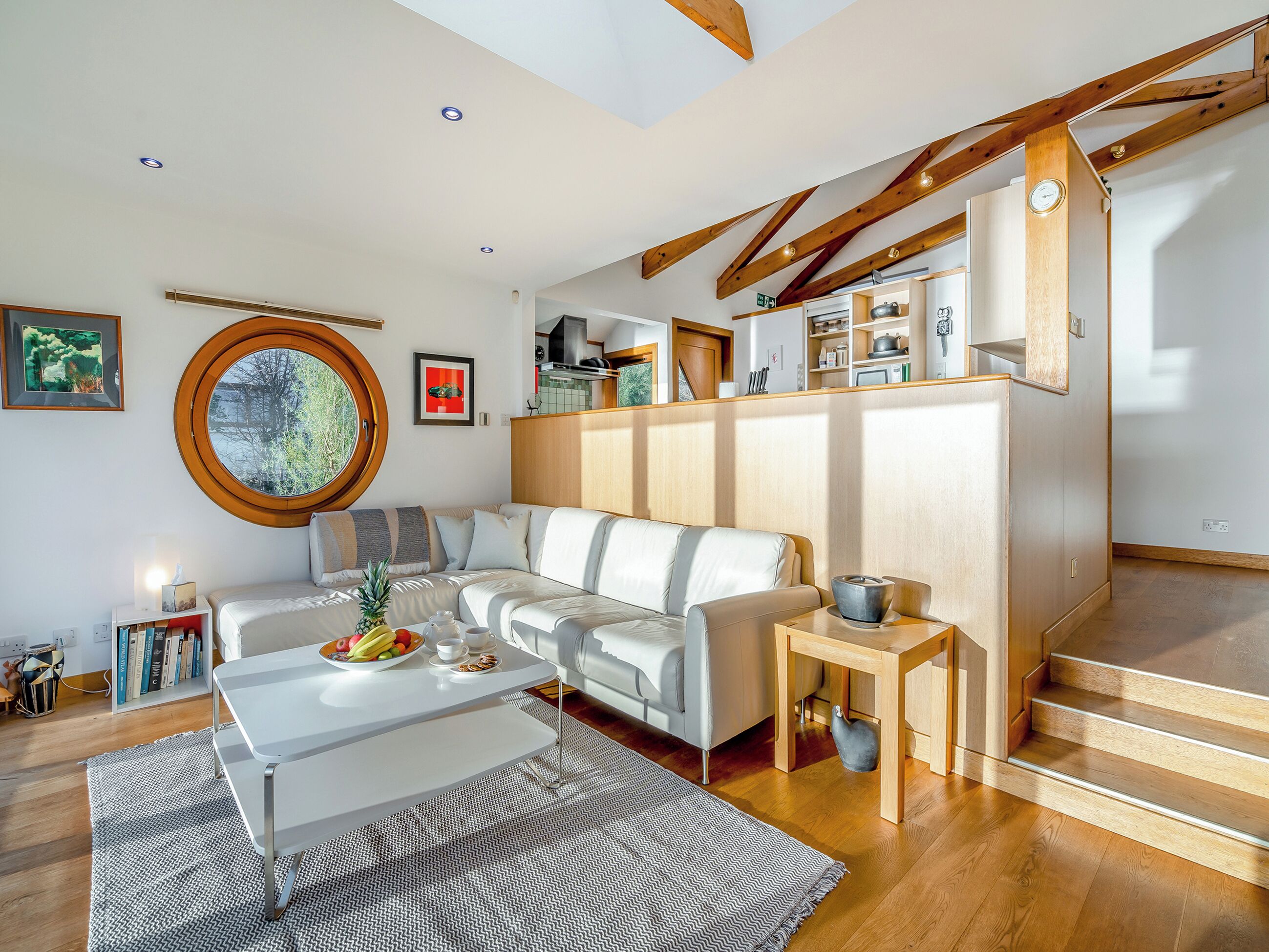 A modern living room with a white sofa, a round window, and a kitchen area with wooden beams.