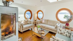A cozy living room with a fireplace, two round windows with views of a lake, a sofa, and a coffee table with a bottle and glasses. - 2 bedroom accommodation in Kilcreggan, near Helensburgh (Kilcreggan, near Helensburgh)