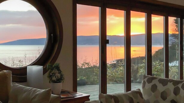 A room with a large window overlooking a body of water and hills during sunset. There is a round mirror on the left and a patterned sofa on the right. - 2 bedroom accommodation in Kilcreggan, near Helensburgh (Kilcreggan, near Helensburgh)