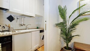 Cuina amb armaris blancs, un rentapeus, una cuina i una màquina de rentar. Hi ha una planta en test i una banca de fusta.