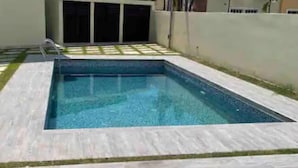 Une piscine rectangulaire dont le bord est recouvert de carrelage, entourée d’une pelouse et d’un mur blanc.