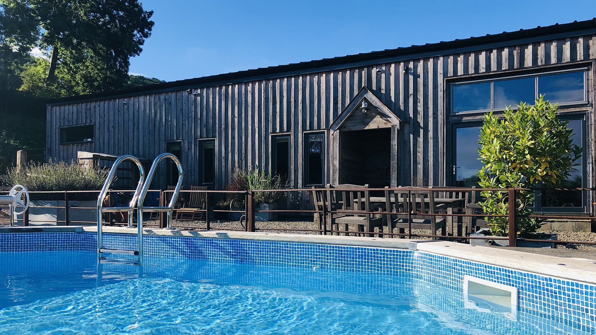 Ein Poolbereich mit einem Holzgebäude im Hintergrund, einer Leiter, die zum Pool führt, und Gartenmöbeln.