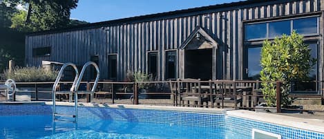 Un espace piscine avec un bâtiment en bois en arrière-plan, une échelle menant à la piscine et des sièges d’extérieur.