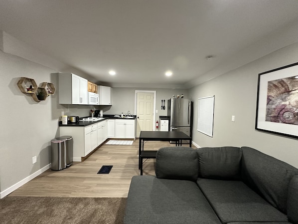 Una cocina moderna con armarios blancos, un refrigerador de acero inoxidable y un comedor con mesa negra y sofá gris.