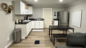 A compact kitchen with white cabinets, a stainless steel refrigerator, and a dining area with a table and couch. - Madison’s Place UNIT B (Peoria)