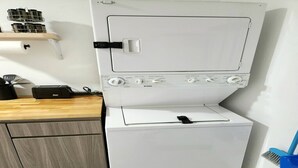 Pieni, valkoinen pyykinpesukone, jossa on ohjauspaneeli. Se sijaitsee pienessä keittiötilassa, jossa on puinen työtaso ja sininen harja.