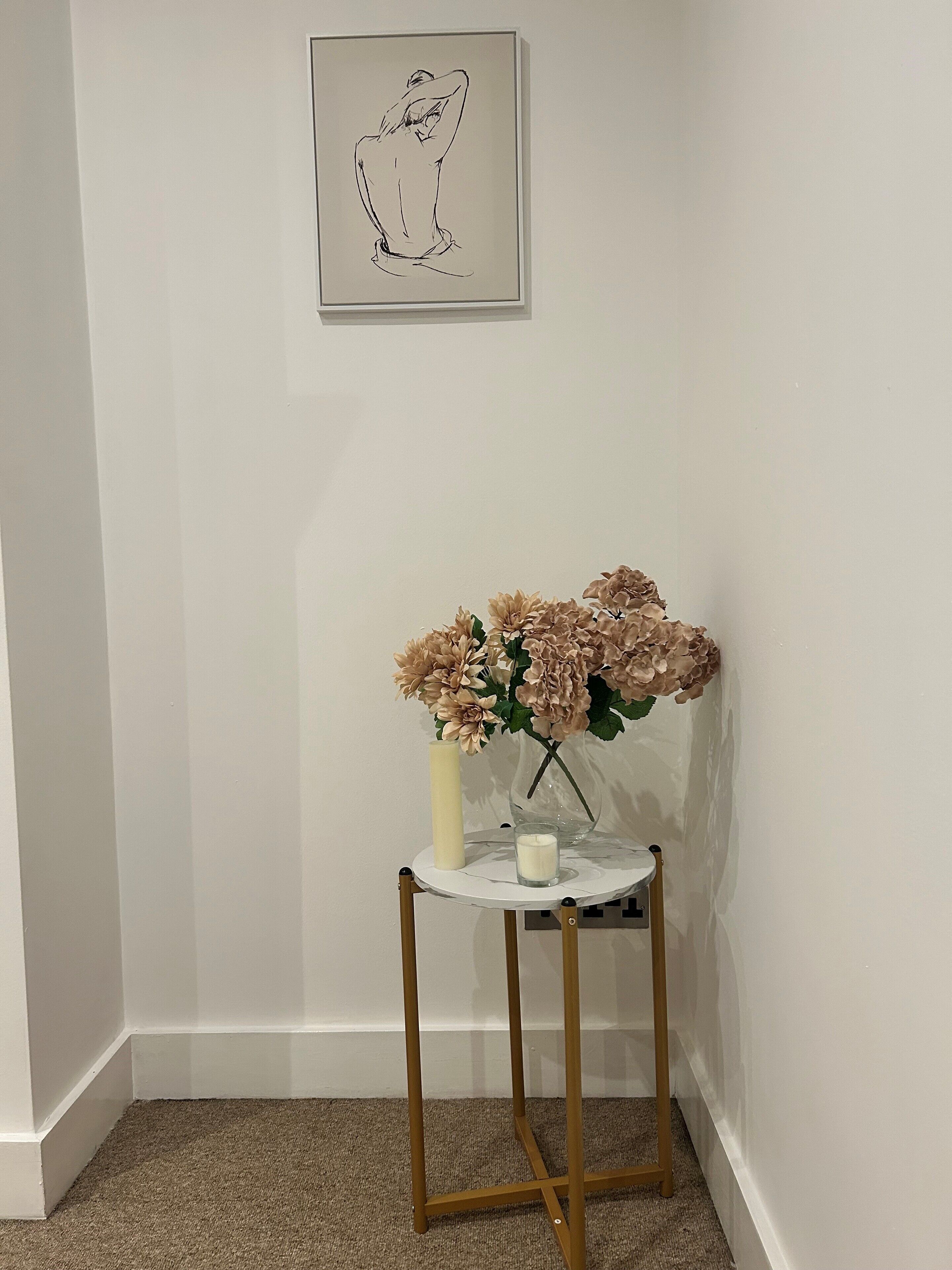 Eine Ecke eines Raumes mit einer weißen Wand, einer gerahmten Zeichnung, einem kleinen runden Tisch mit Blumen und einer Kerze.