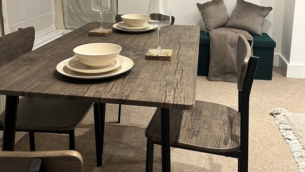 Un comedor con una mesa de madera dispuesta para dos personas, dos sillas, un sofá verde y un estante en la pared con objetos decorativos.