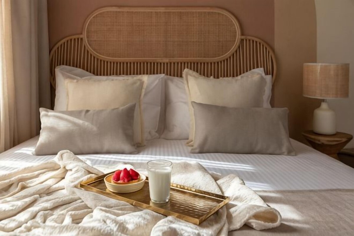 Una cama con edredón blanco, una bandeja con un cuenco de fruta y un vaso de leche, y una lámpara de noche.
