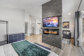 A modern bedroom with a large bed, a fireplace, a mounted TV, and a view of the living area. - Mountain Escape With Fireplace (Show Low)