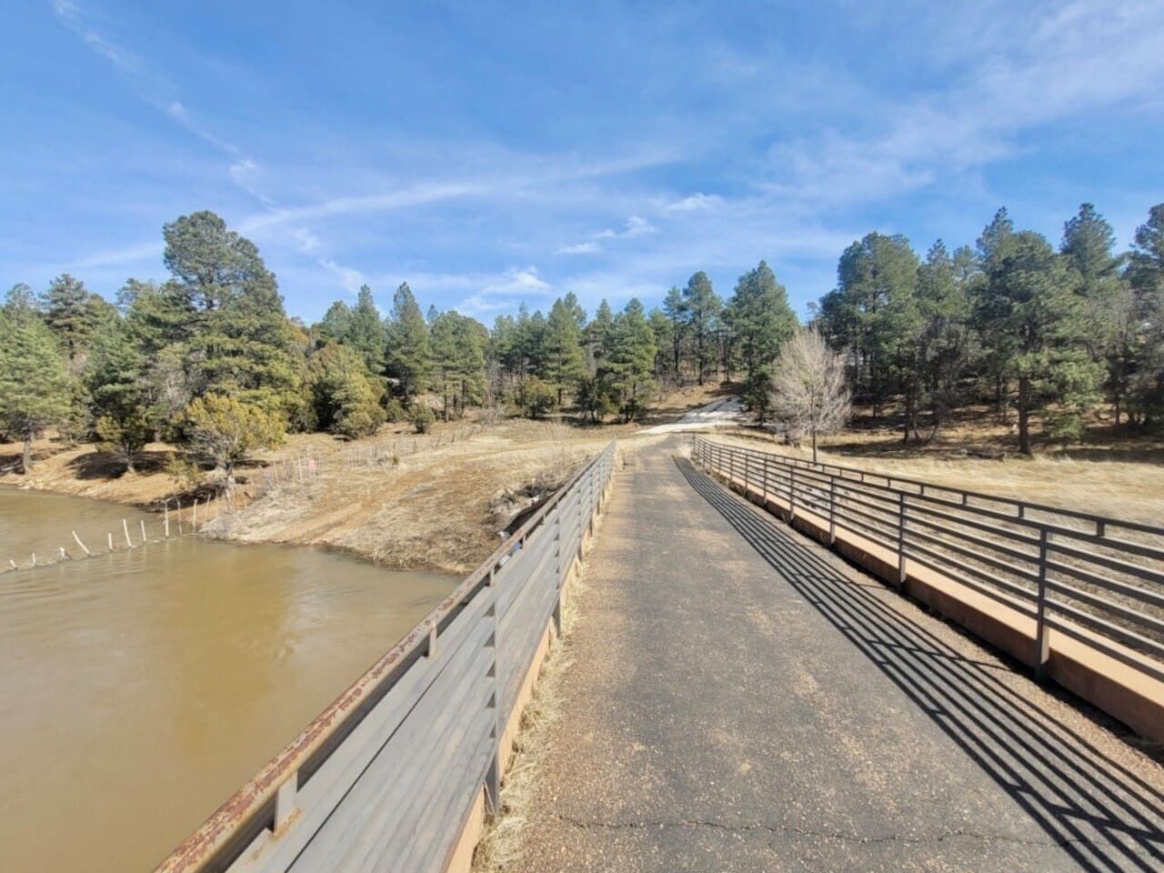 A bridge with a metal railing, a dirt path, and a body of water on one side.