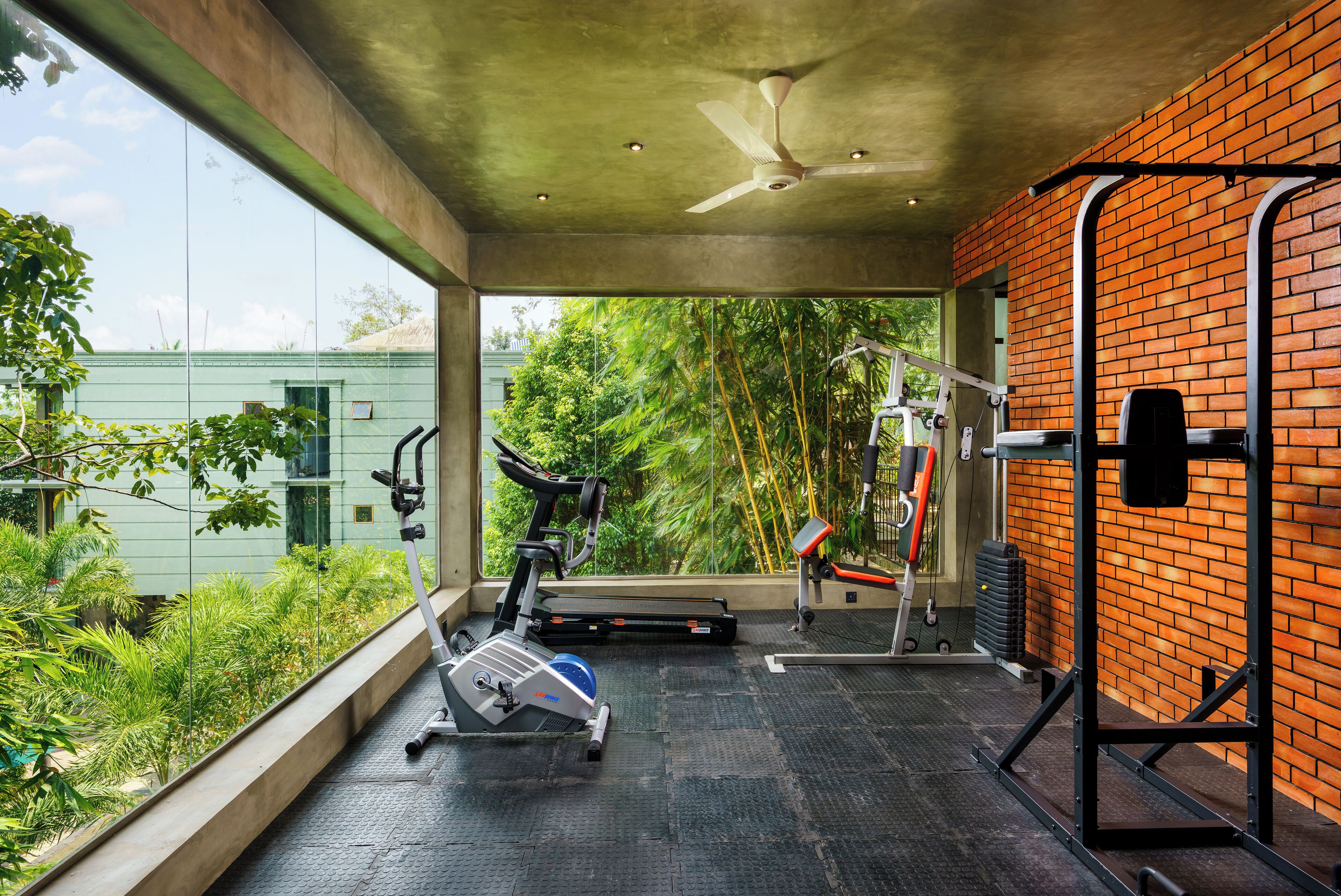 Un gimnasio con equipo de ejercicio, ventilador de techo y vista a zonas verdes al exterior.