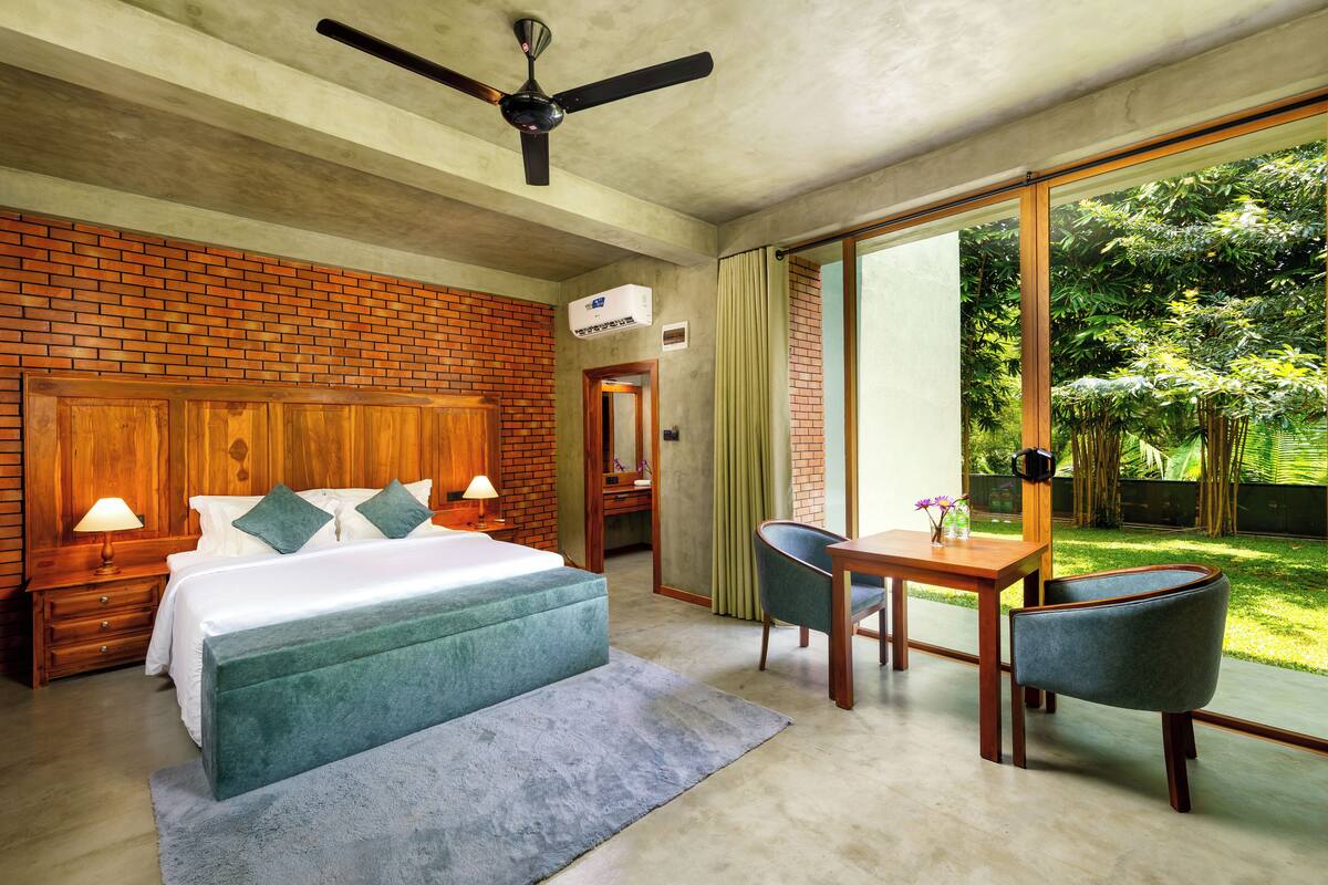 Une chambre moderne avec un mur en briques, un grand lit, un ventilateur de plafond et une vue sur un jardin luxuriant, visible à travers une porte-fenêtre coulissante.