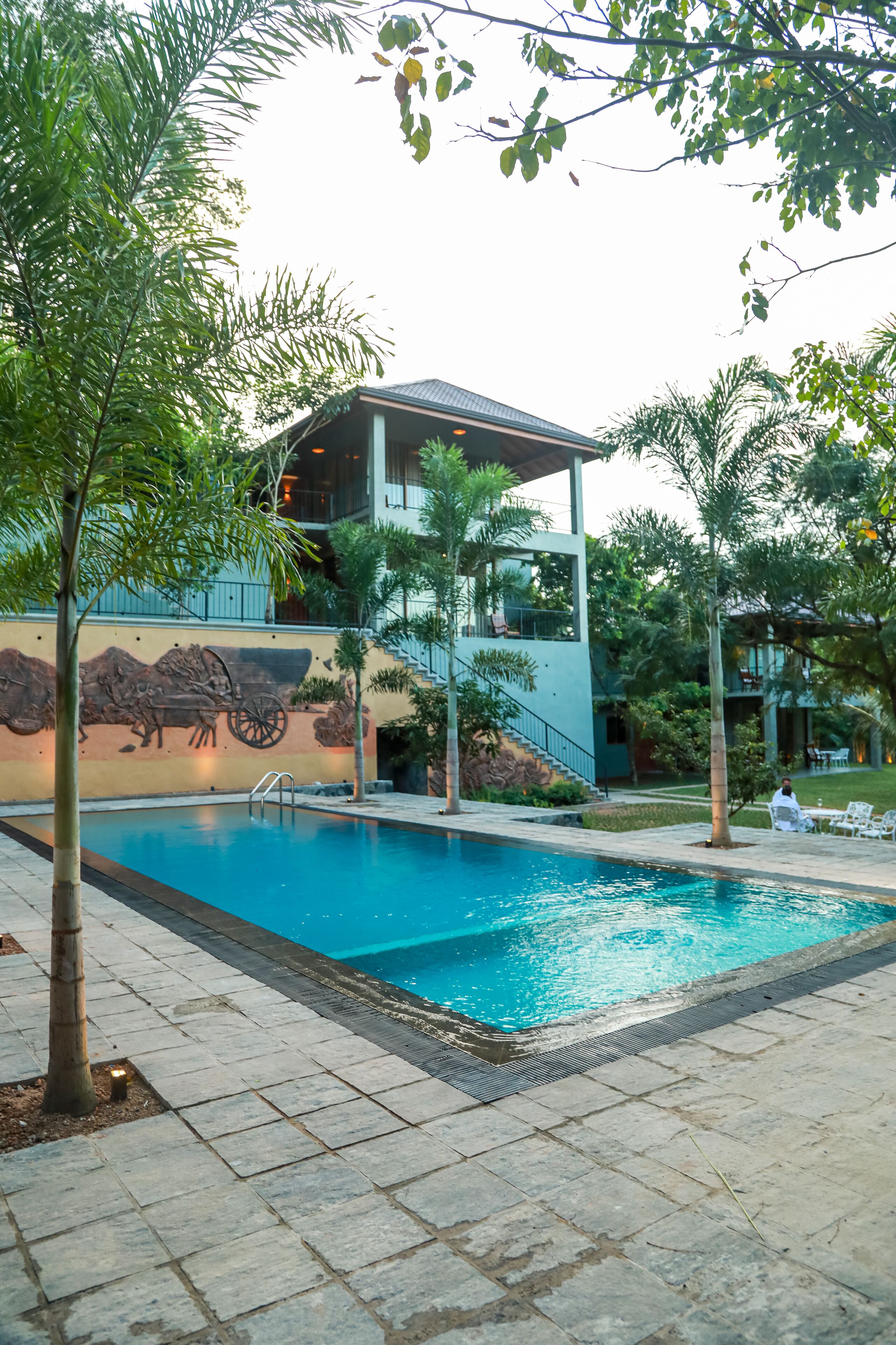 A swimming pool with a stone border, surrounded by palm trees and a building with a mural.