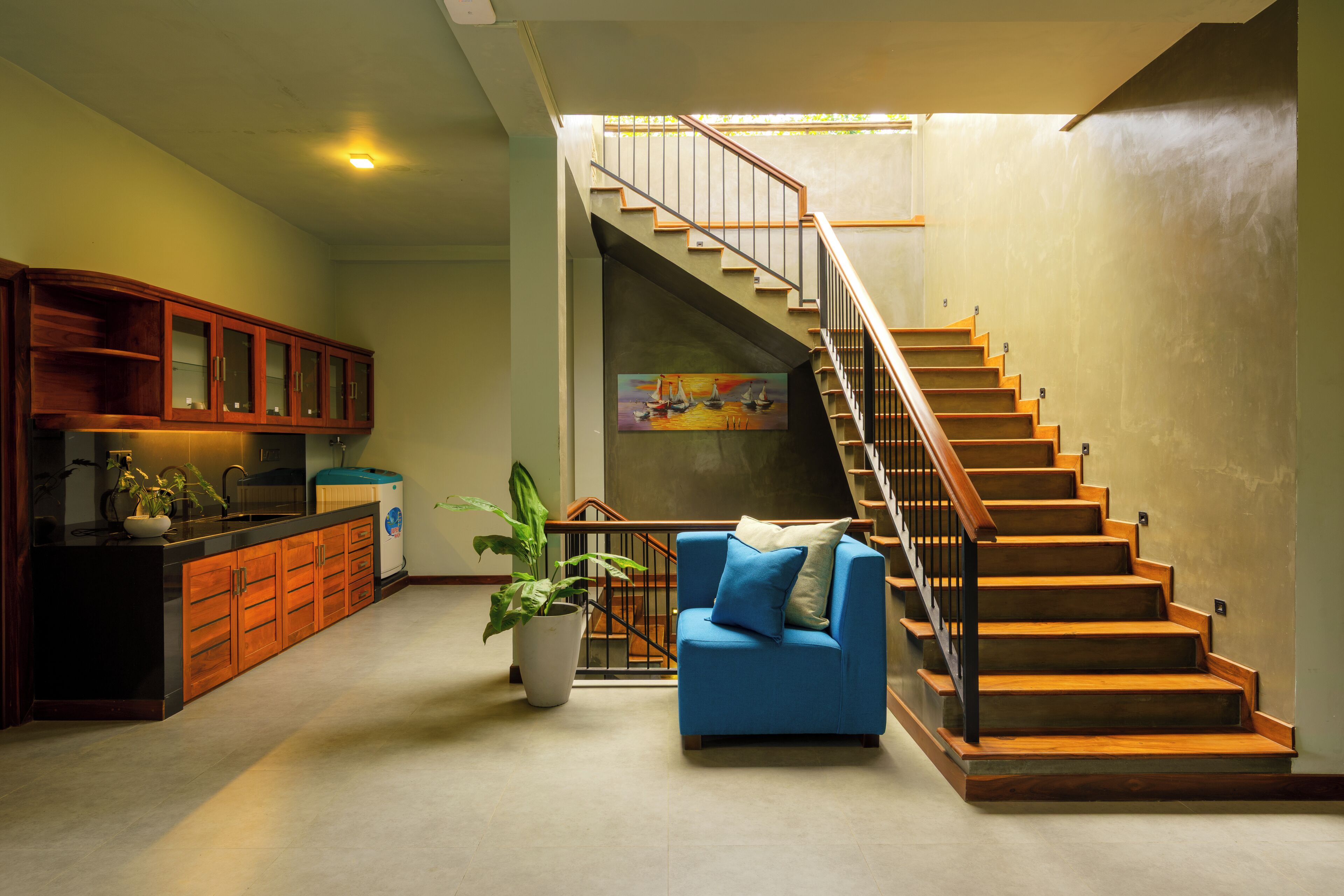 A spacious living area with a kitchen, a blue sofa, a staircase, and a painting on the wall.