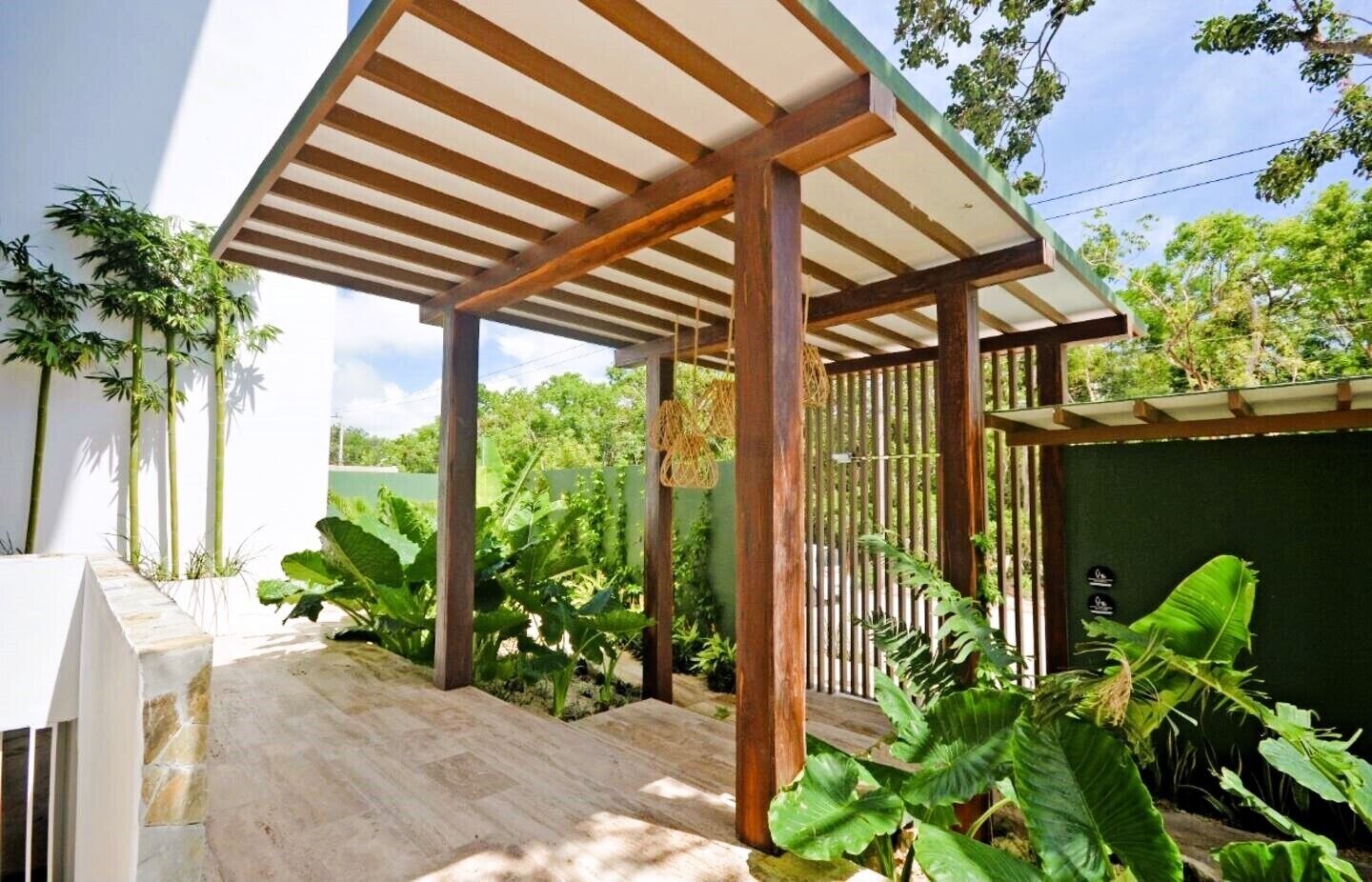 A covered outdoor area with a wooden pergola, stone flooring, and lush greenery.