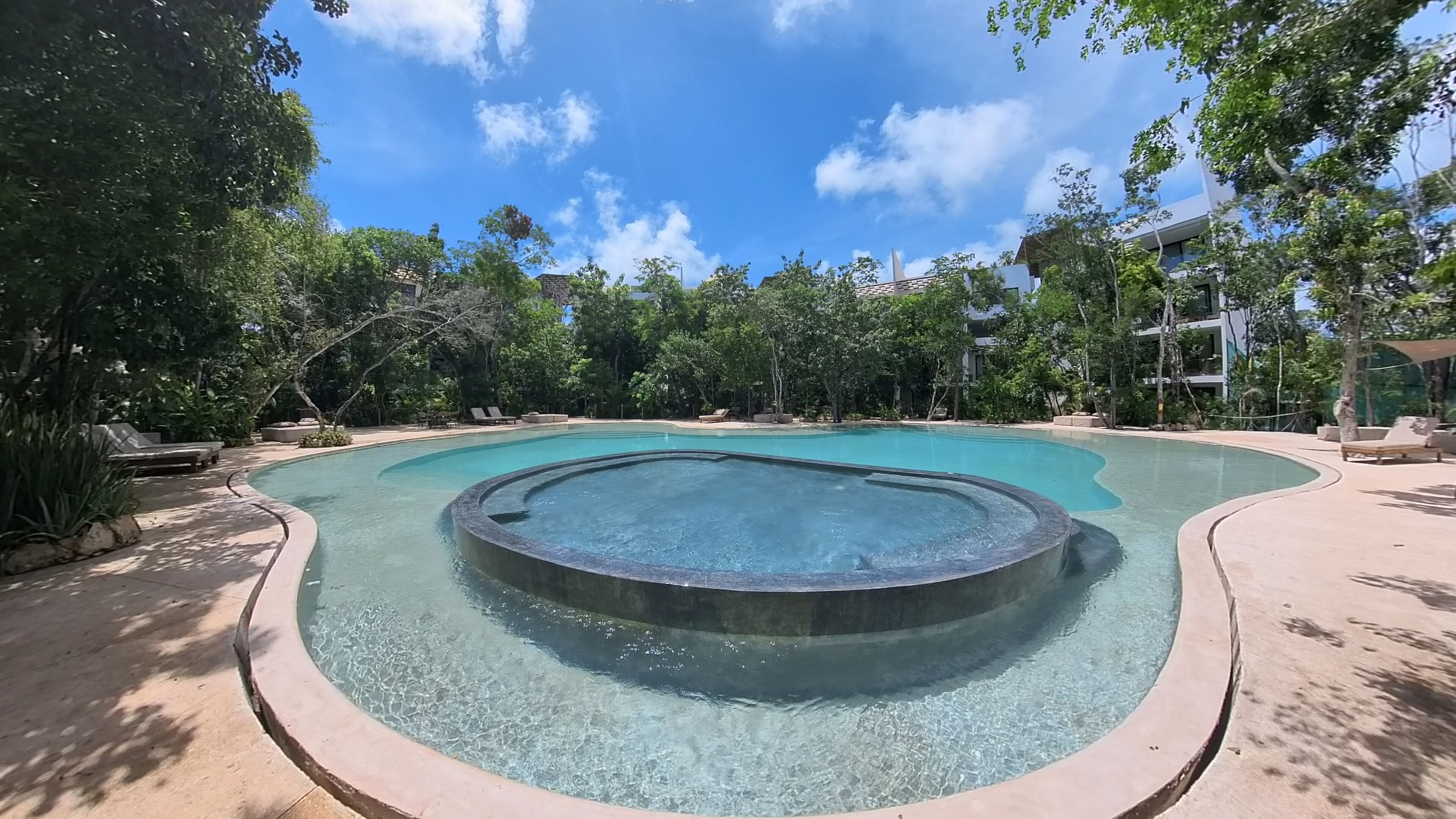 Une grande piscine extérieure avec un jacuzzi au centre, entourée de chaises longues et d’arbres.