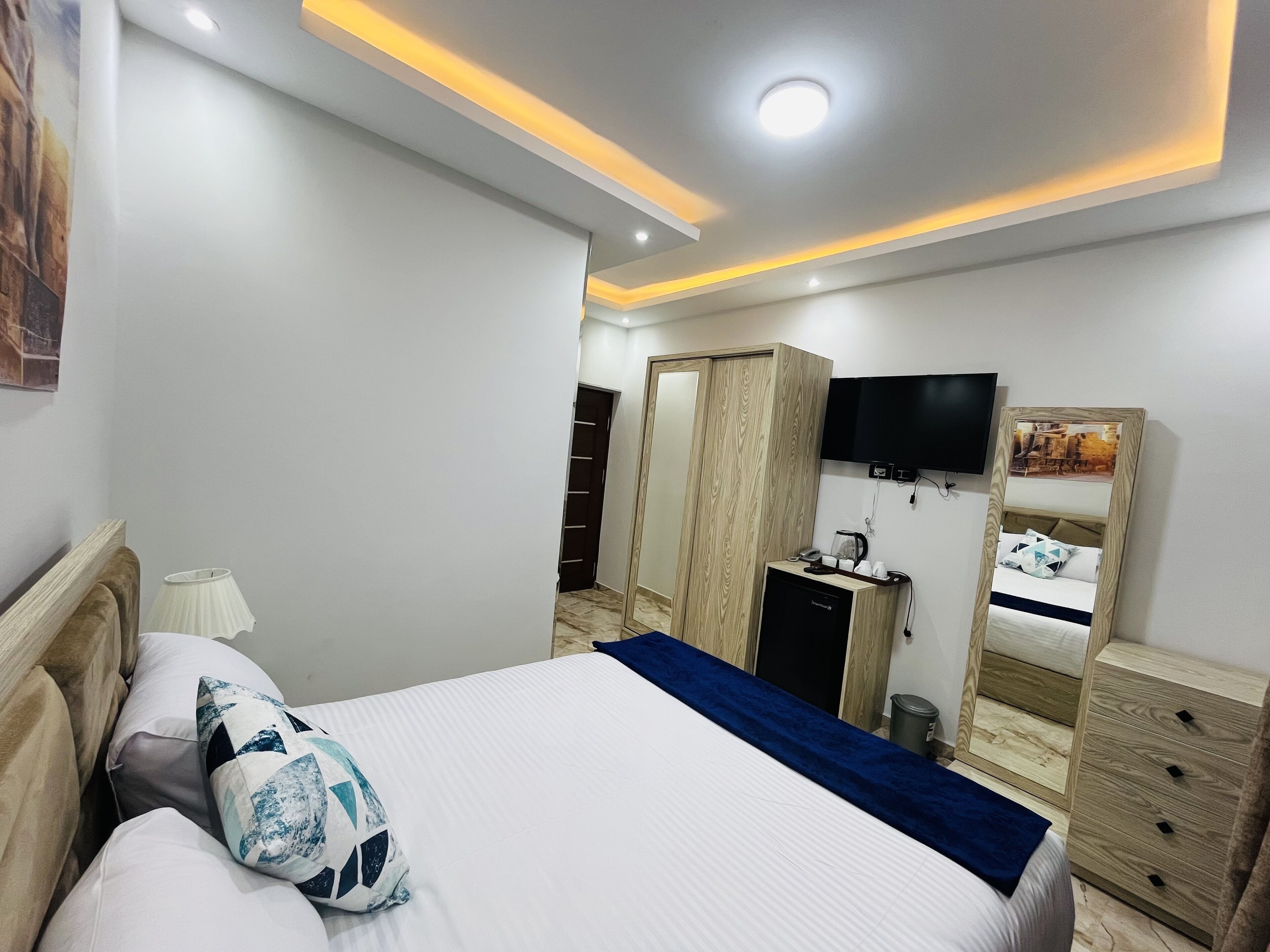 Ein modernes Hotelzimmer mit Bett, Nachttisch, Lampe, Kleiderschrank und einem an der Wand befestigten Fernseher.