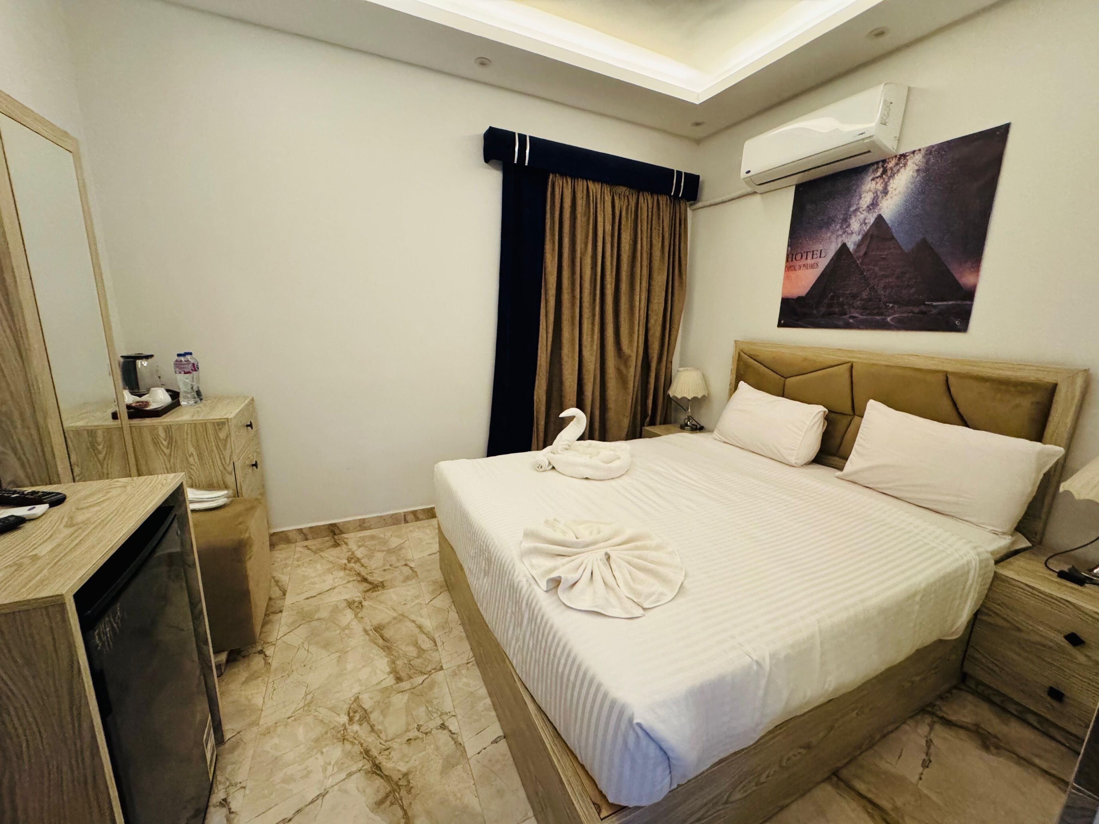 A hotel room with a bed, a nightstand, a desk, a mirror, a wall-mounted air conditioner, and a poster of the pyramids.