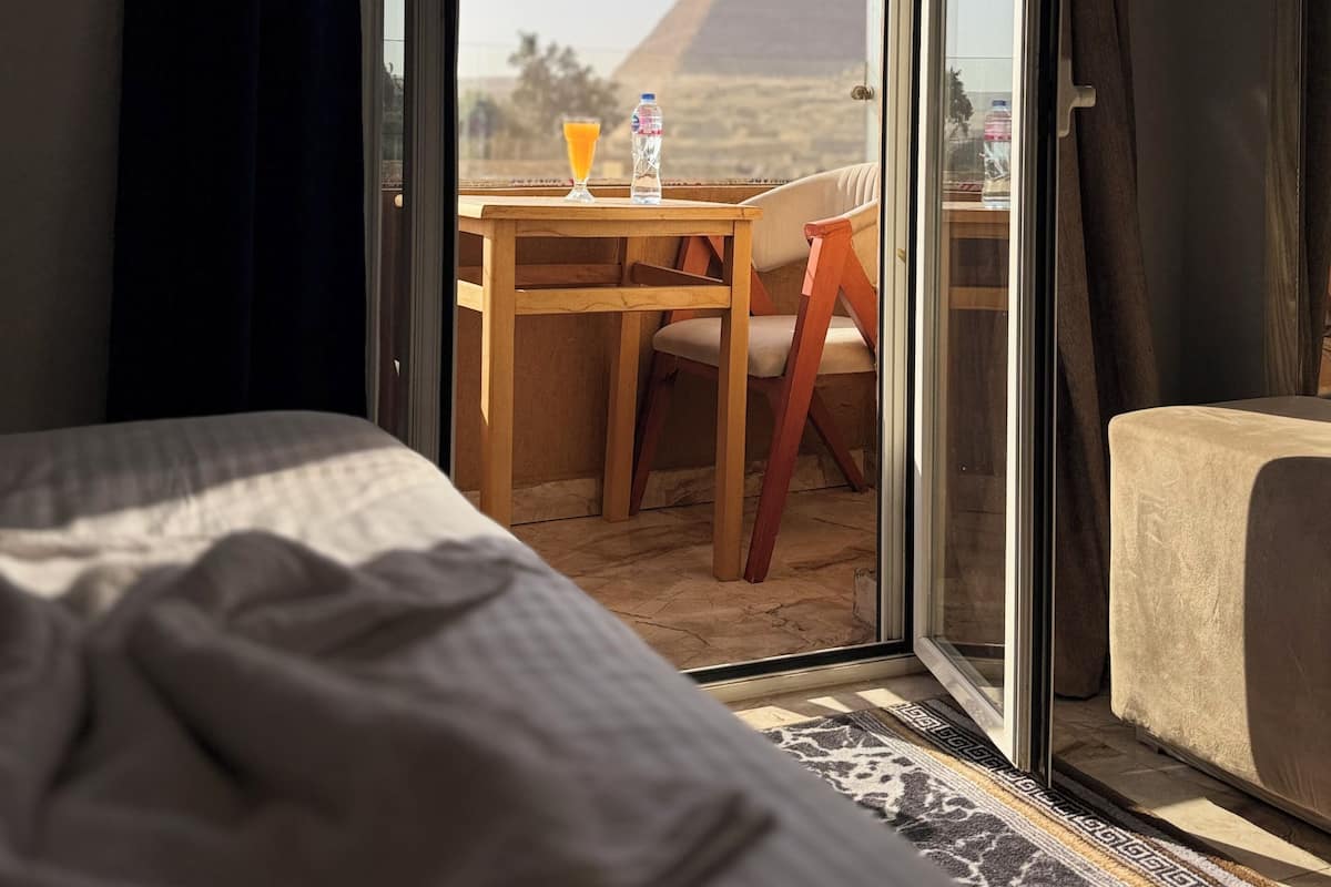 Et hotelværelse med en seng, et bord med en stol og udsigt til en pyramide gennem en glasdør.