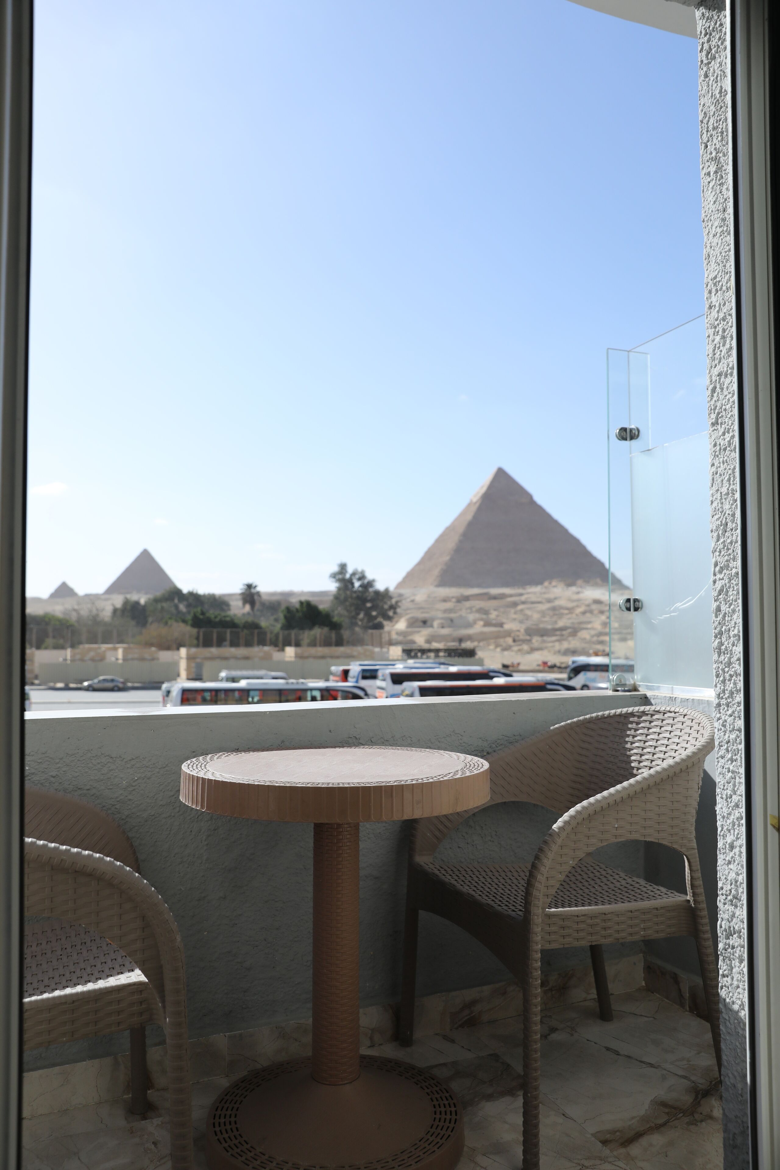 A balcony with wicker chairs and a round table, overlooking a parking area and pyramids in the background.
