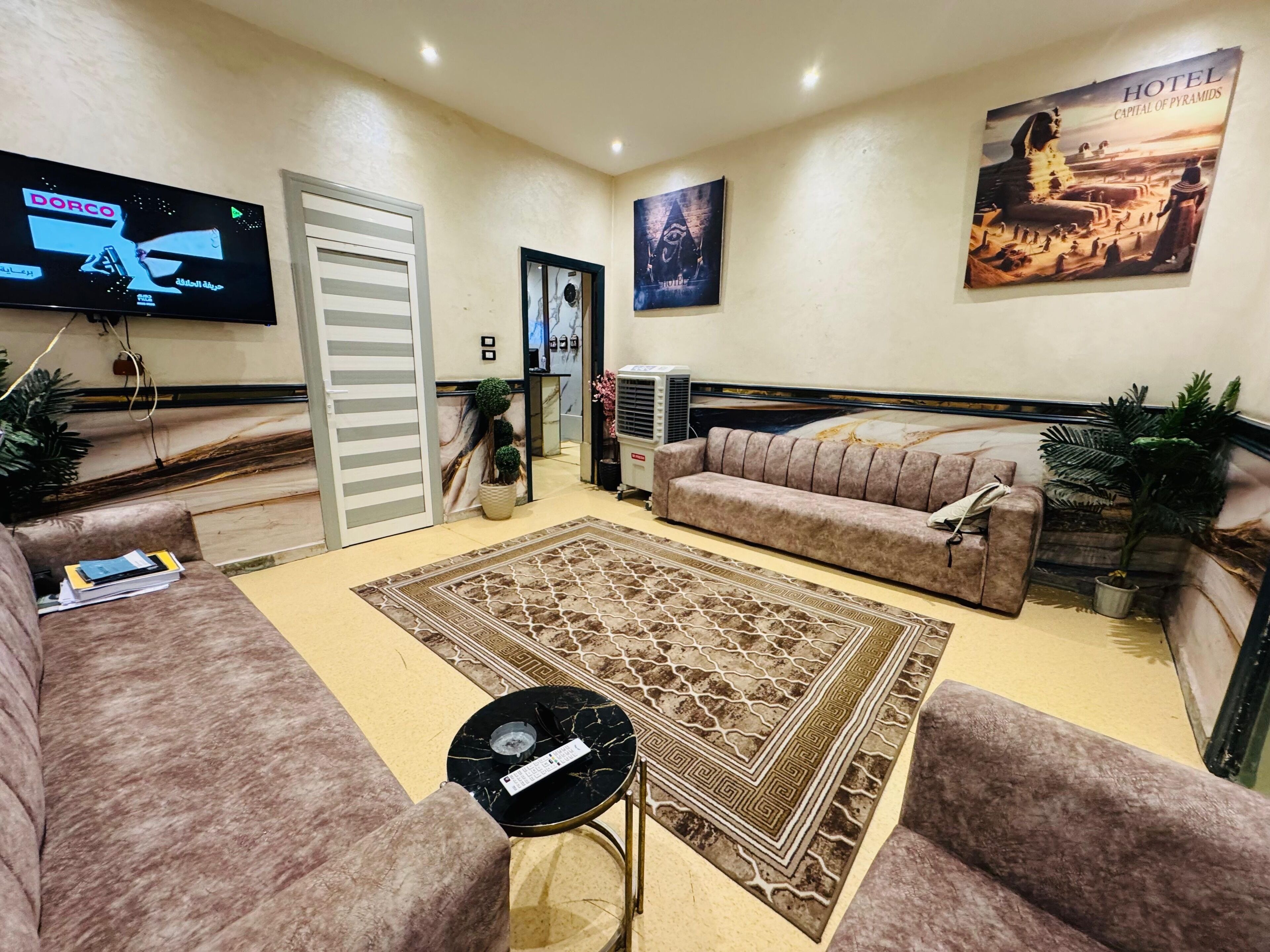 A hotel room with a large bed, two sofas, a coffee table, a TV, a painting of the pyramids, and a potted plant.