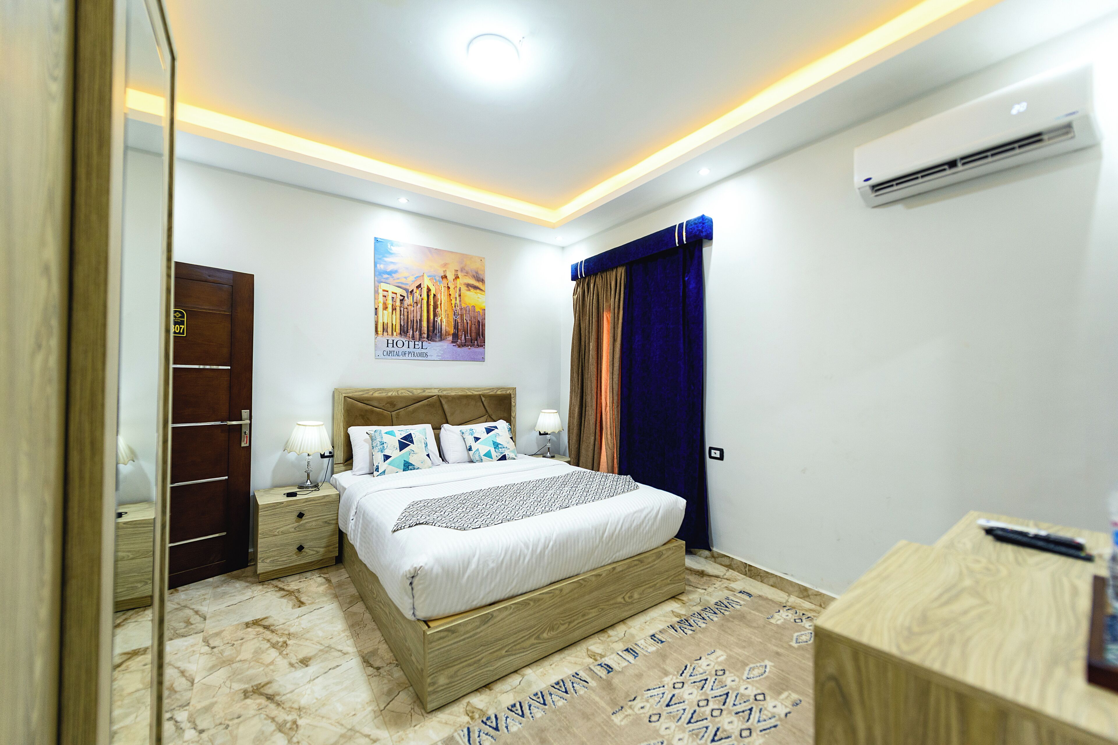 A hotel room with a bed, bedside tables, a mirror, an air conditioner, and a poster on the wall.