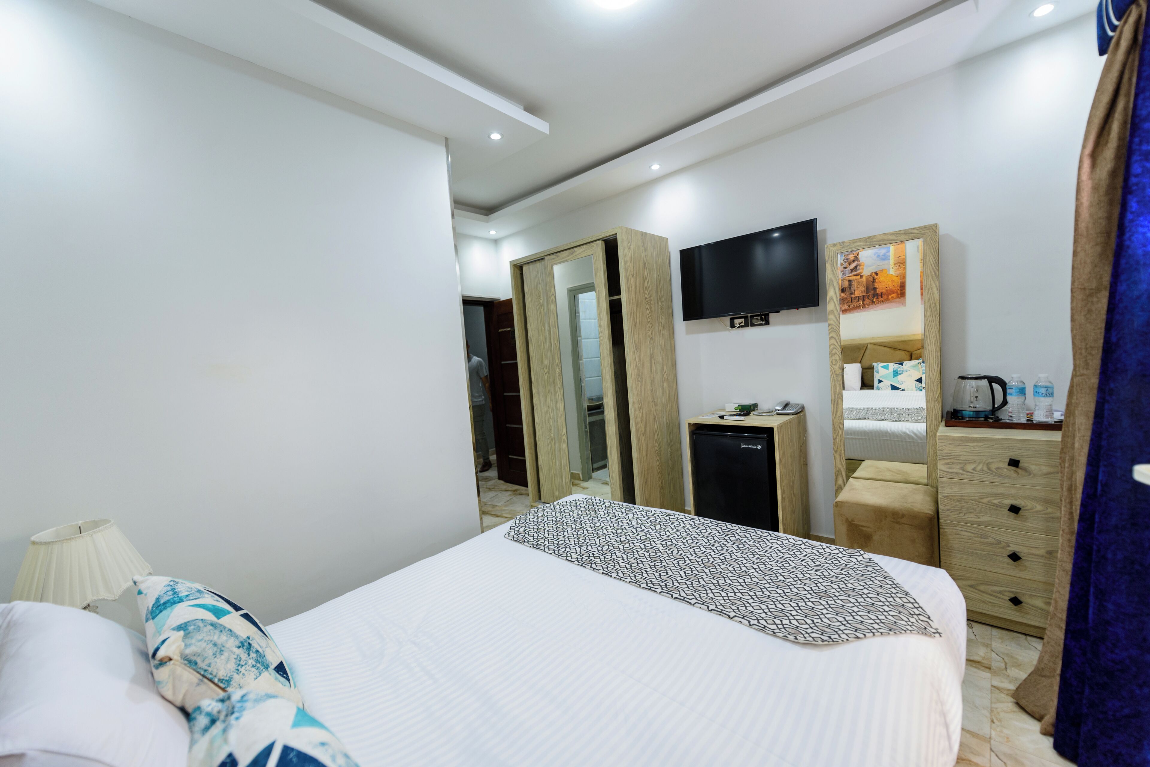 Habitación de hotel con cama, televisión, espejo, cómoda y escritorio con tetera.