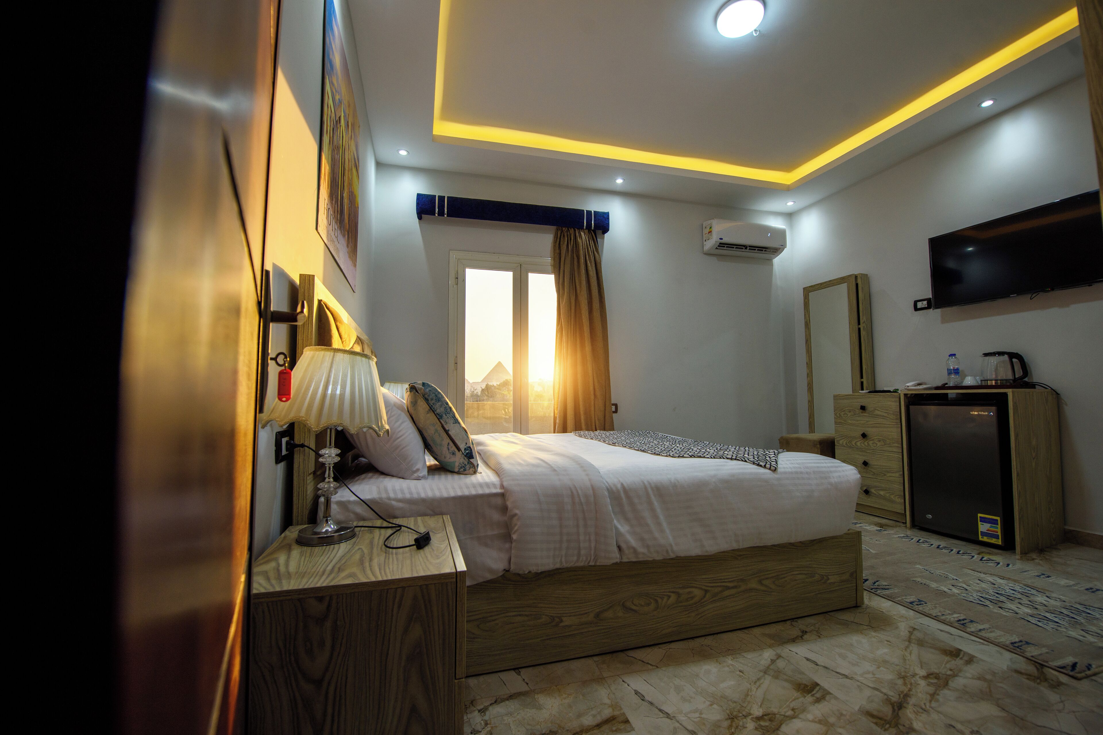Una habitación de hotel moderna con cama, mesita de noche, lámpara, aire acondicionado, televisor y un pequeño refrigerador.