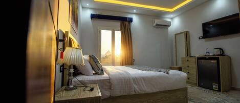 Una habitación de hotel moderna con cama, mesita de noche, lámpara, aire acondicionado, televisor y un pequeño refrigerador.