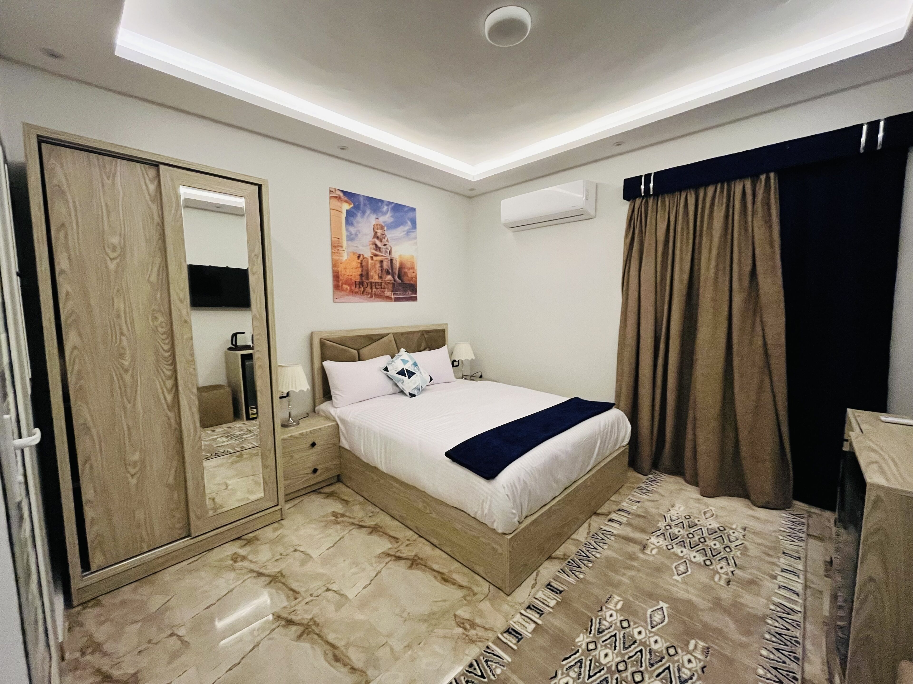 A hotel room with a bed, a wardrobe, a mirror, an air conditioning unit, and a patterned rug.