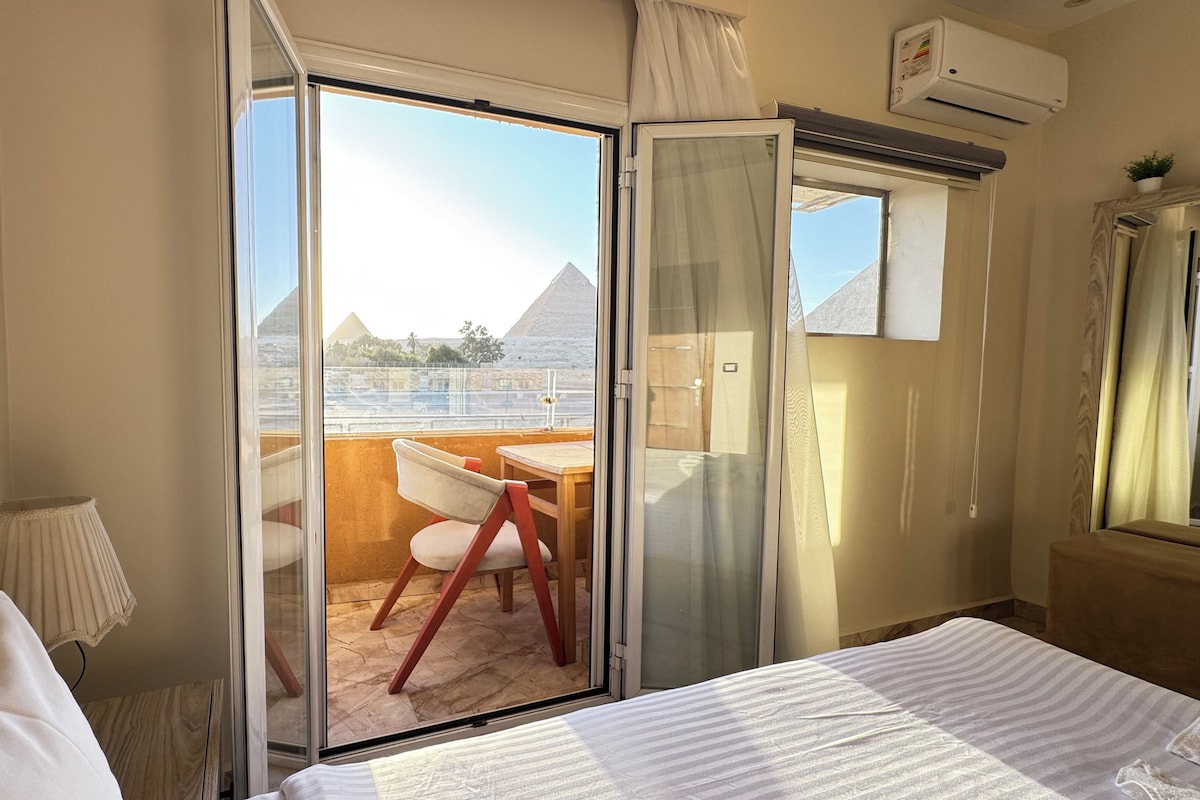 Habitación de hotel con cama, una silla, una mesa y puerta corrediza de vidrio que da a un balcón con vista a una pirámide.