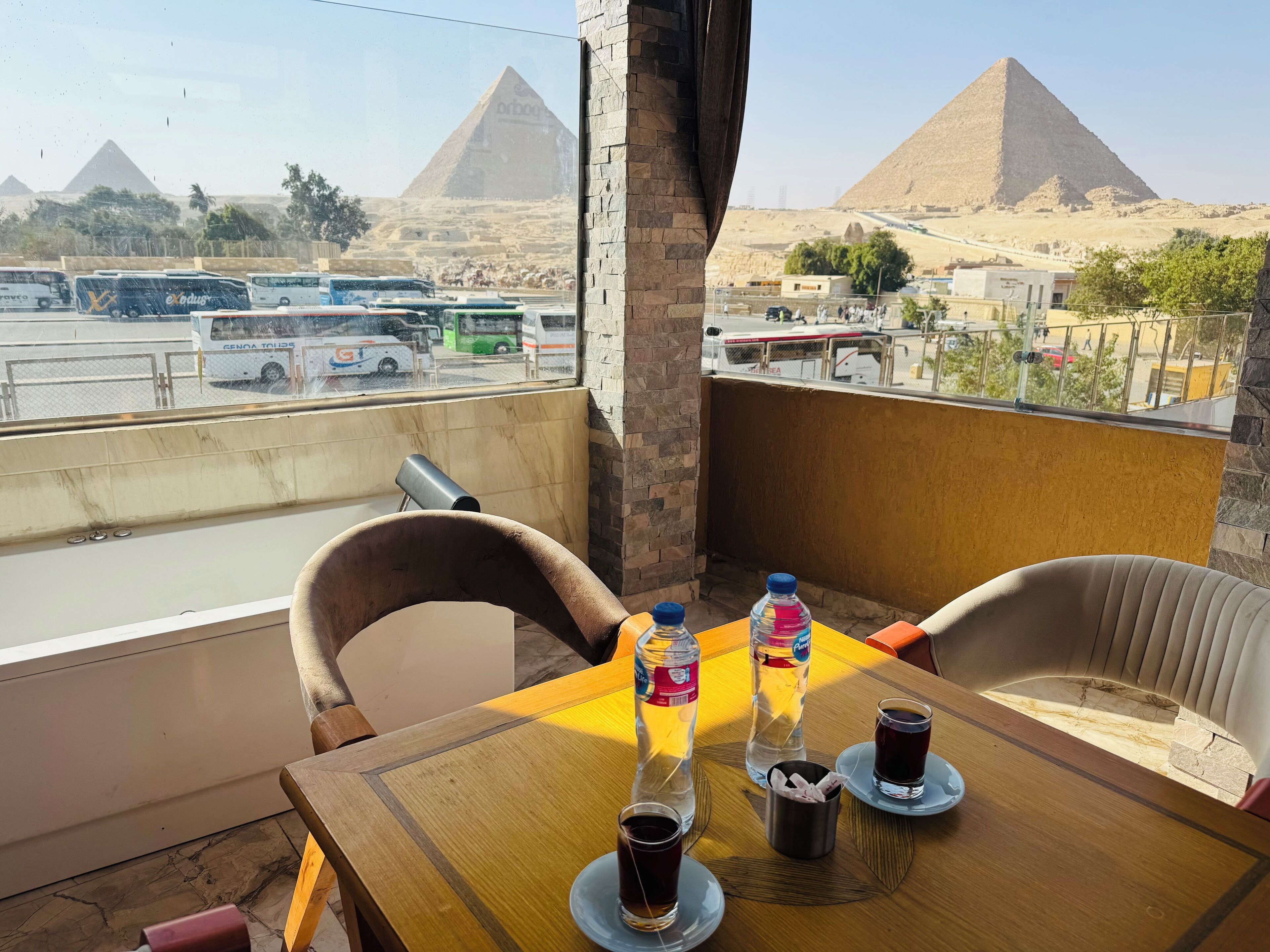 A balcony with a table set for two, featuring two bottles of water, a cup of tea, and a small metal cup. The view includes pyramids and a parking area with buses.