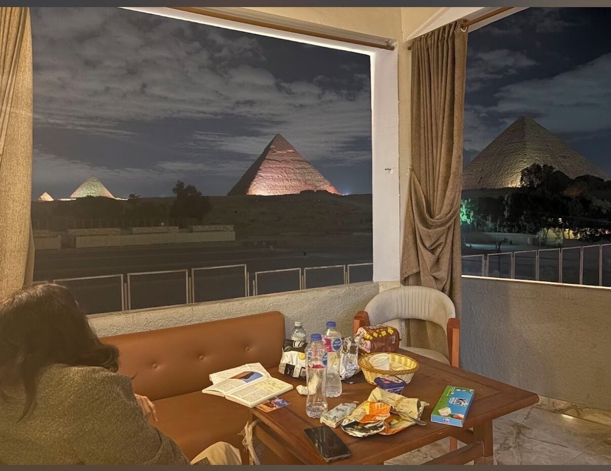 A balcony with a view of the pyramids at night, a sofa, a table with snacks and drinks, and a person reading.