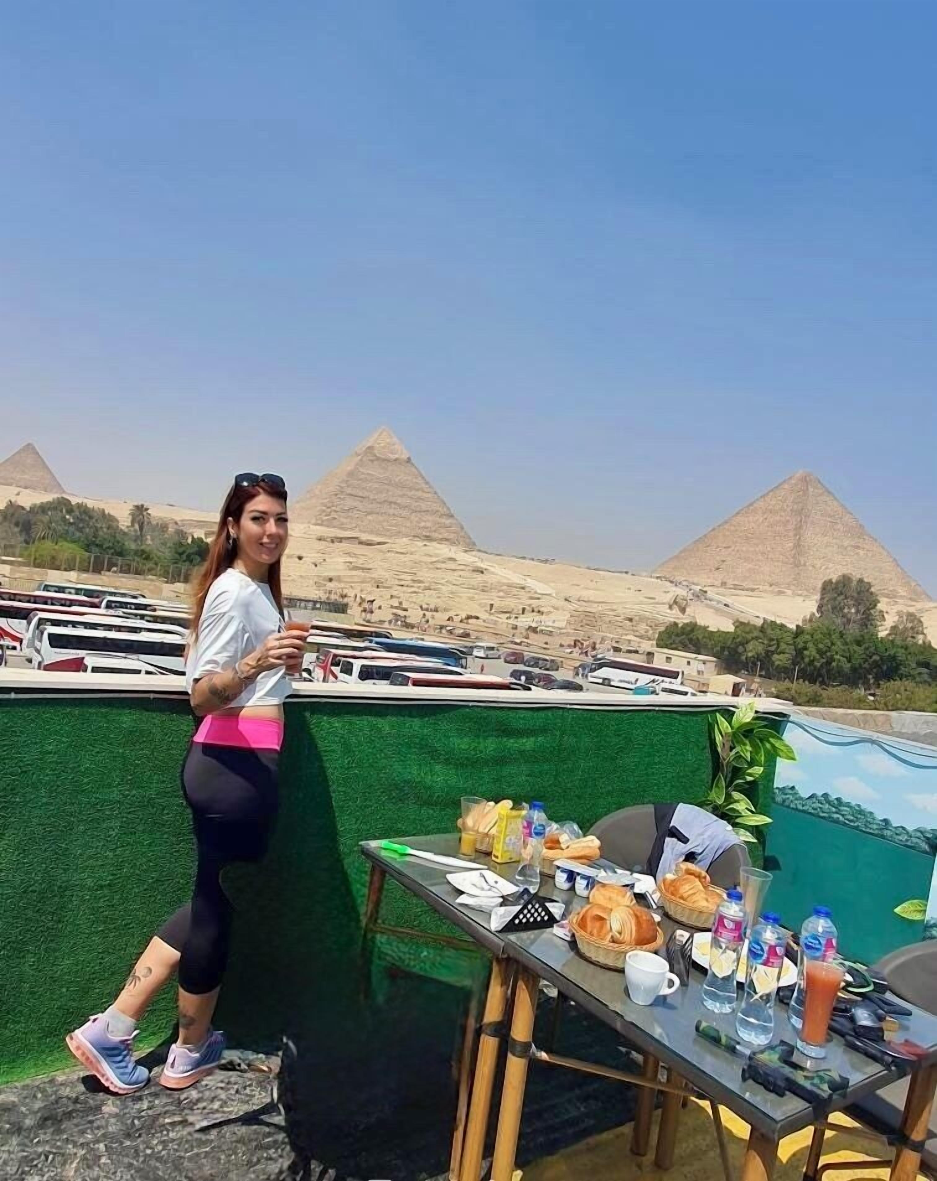 A person standing by a table with food and drinks, overlooking a view of the pyramids.