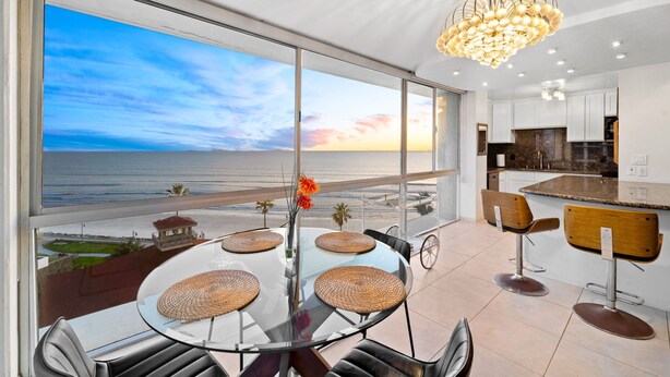 A modern kitchen with a view of the ocean, a dining area with a glass table and chairs, and a chandelier.