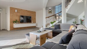 A modern living room with a flat-screen TV, wooden panel wall, grey sofa, staircase with decorative lighting, and a white coffee table.