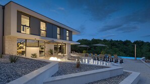 A modern two-story house with a well-lit outdoor area featuring a chessboard, lounge chairs, and a swimming pool.