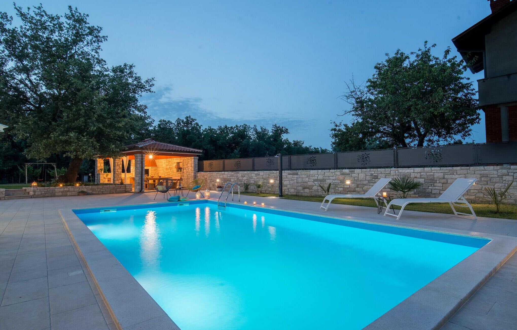 Una piscina con sillas de descanso, un muro de piedra y un pequeño pabellón con luces.