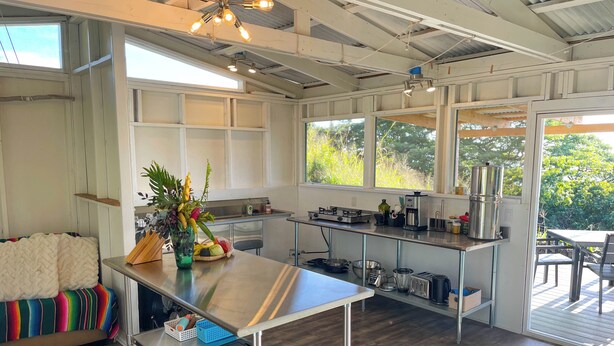 A modern kitchen with stainless steel appliances, a dining area with a view, and a cozy seating area with a colorful blanket. - Pueo Cabin with Shared Bath and Common House (Kealakekua)