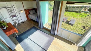 A room with a bed, a small table with a plant, a metal shelf unit, and a view of the outdoors. - Pueo Cabin with Shared Bath and Common House (Kealakekua)