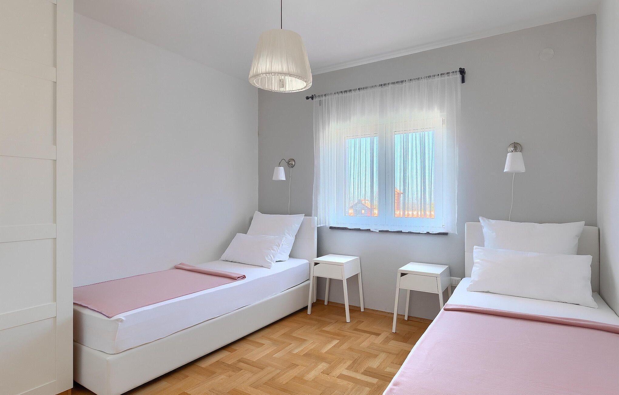 A bedroom with two single beds, a small bedside table, a nightstand, a window with sheer curtains, and a hanging light fixture.
