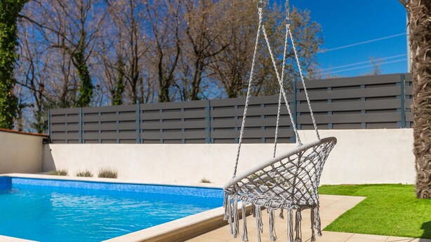 Un fauteuil suspendu près d’une piscine, avec vue sur une clôture et des arbres.