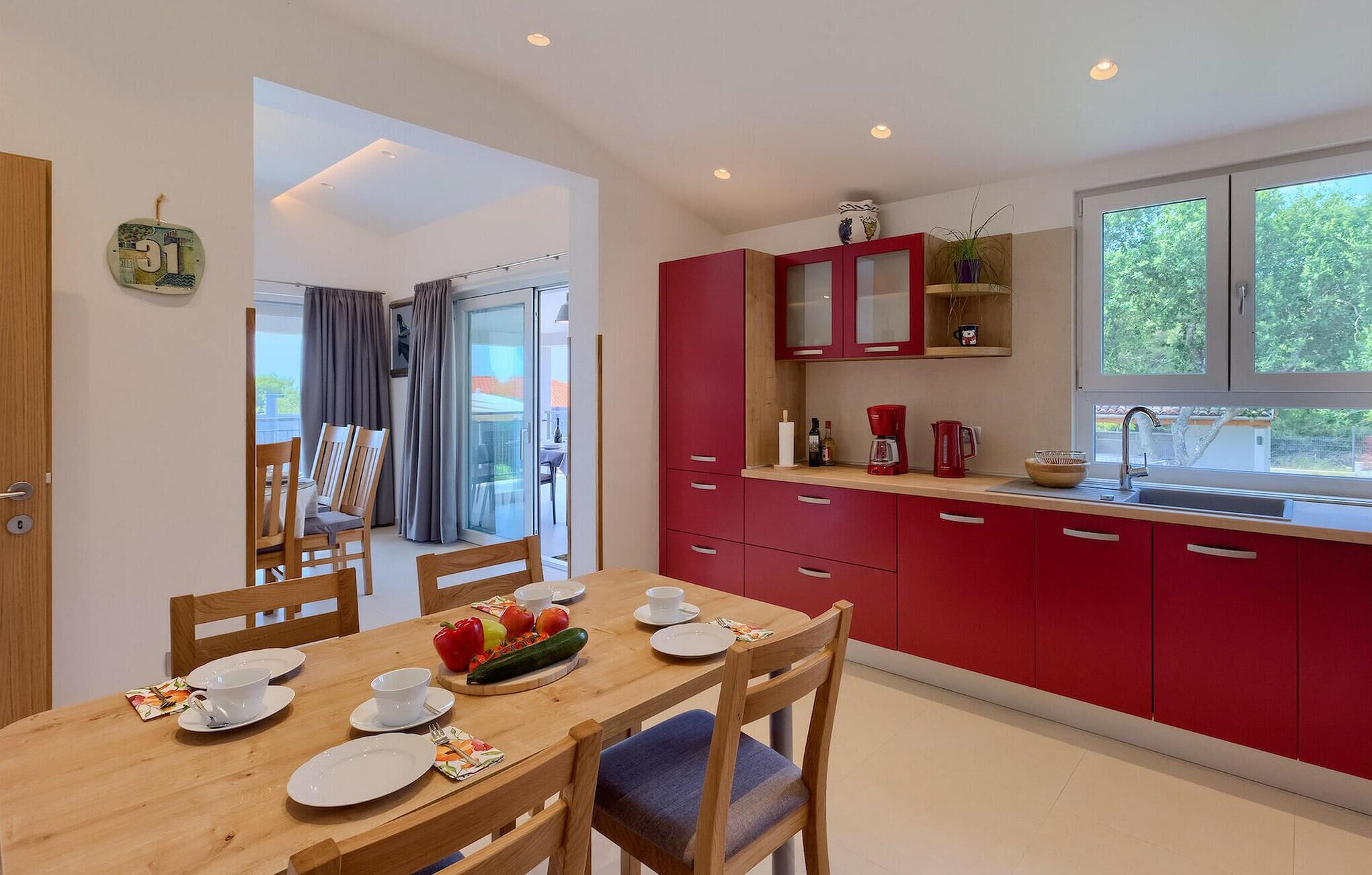 A modern kitchen with red cabinets, a wooden dining table with chairs, and a window with a view of greenery.