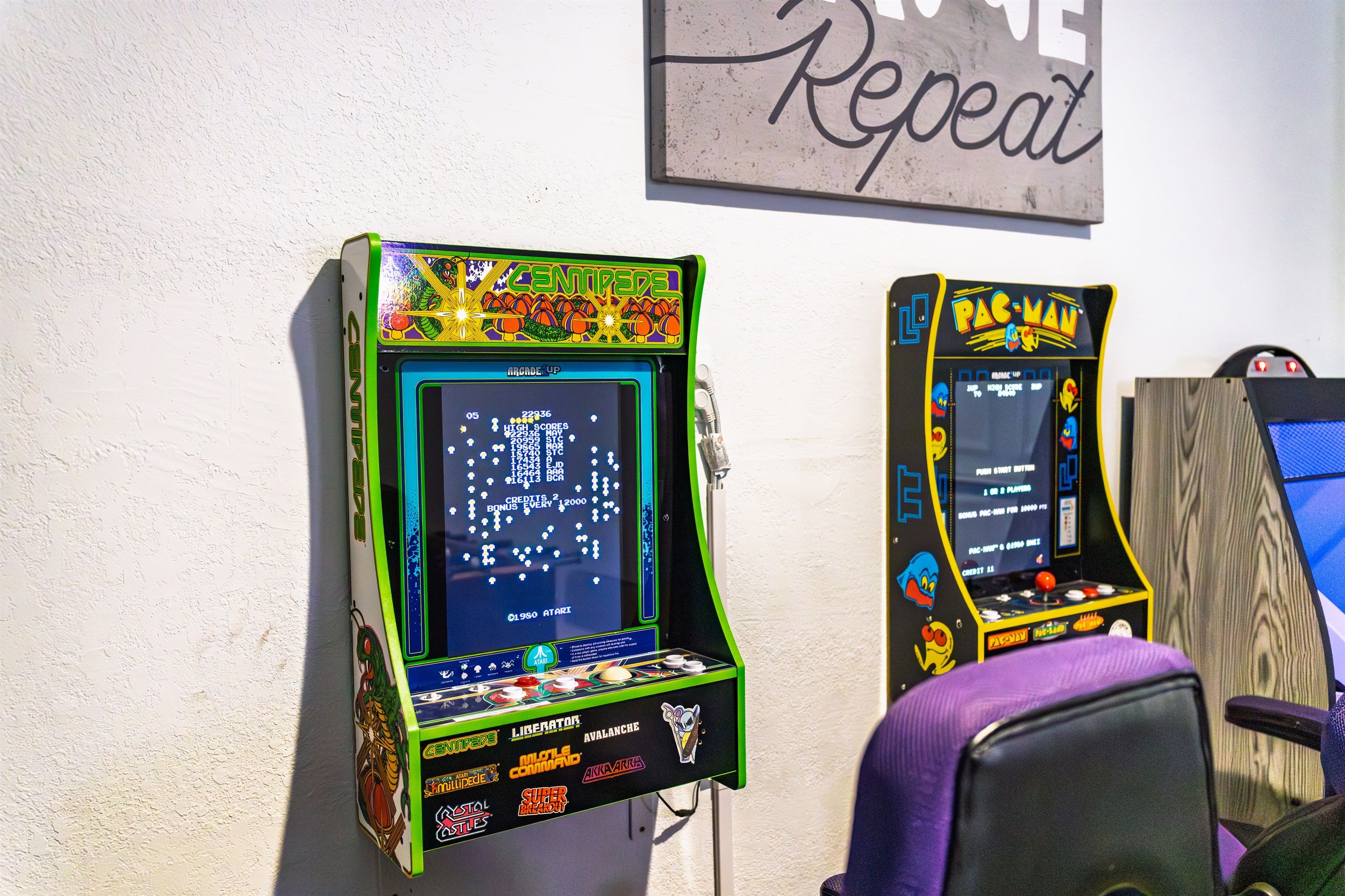 Two arcade machines, one labeled 'Centipede' and the other 'Pac-Man', mounted on a wall with a sign above them that reads 'Repeat'.