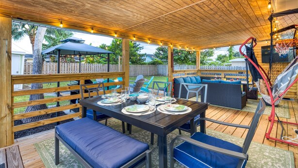 An outdoor patio with a wooden deck, a dining table set with plates and glasses, blue cushioned seating, and a basketball hoop. - Arcade · Firepit · 2 King Beds + Workspace (Stuart)