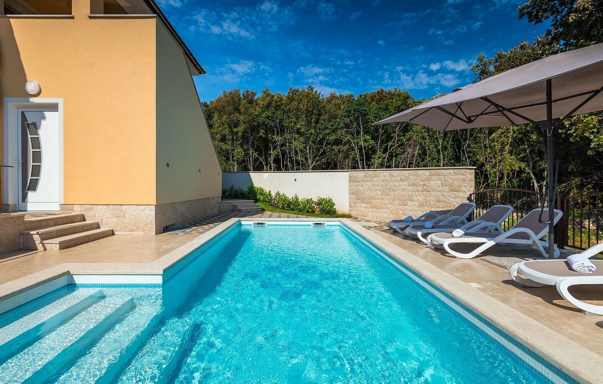A swimming pool with clear blue water, surrounded by a beige building with a door and steps leading to the pool. There are lounge chairs and a large umbrella providing shade.